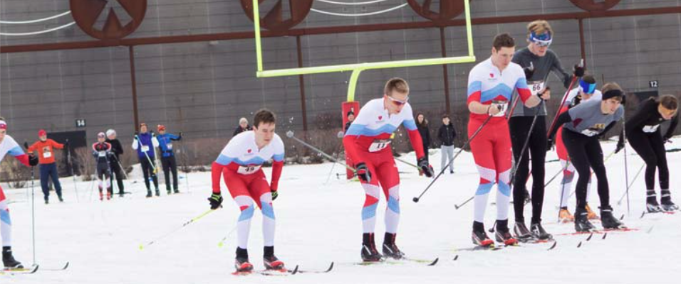 A group of Nordic skiers at the start of a race on a snowy field. Some athletes are in red, white, and blue suits, while others wear dark-colored gear. Spectators and officials watch from the background, near an outdoor sports structure.